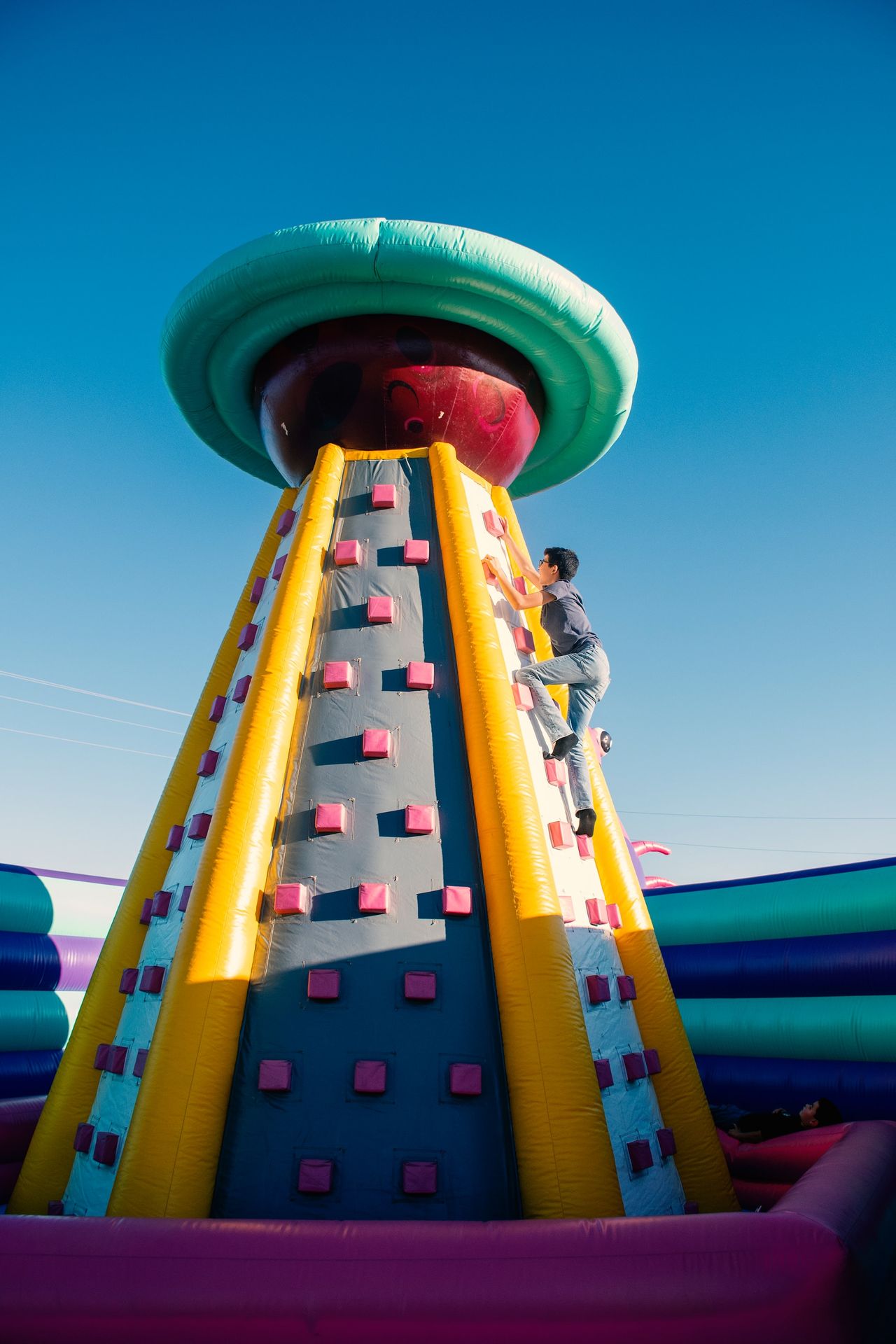nflatable Climbing Wall with Mushroom Top – Outdoor Fun Attraction A tall inflatable climbing structure with blue and gray climbing wall panels, yellow side rails, pink footholds, and a large green rotating mushroom-like top against a clear blue sky; children are climbing the wall.