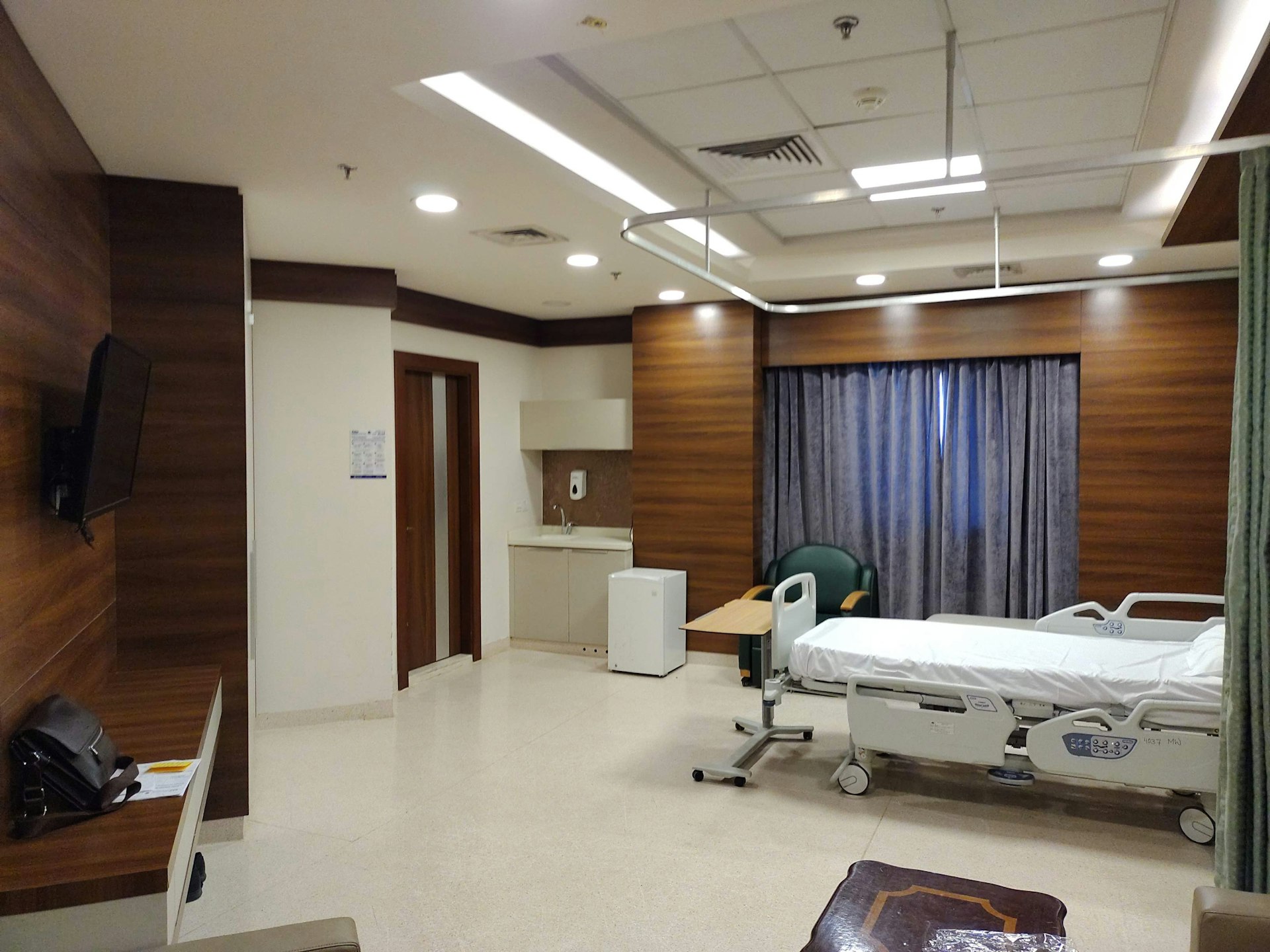 A well-appointed hospital room featuring a patient bed, a TV mounted on the wall, a small kitchenette, and a seating area with a green chair.