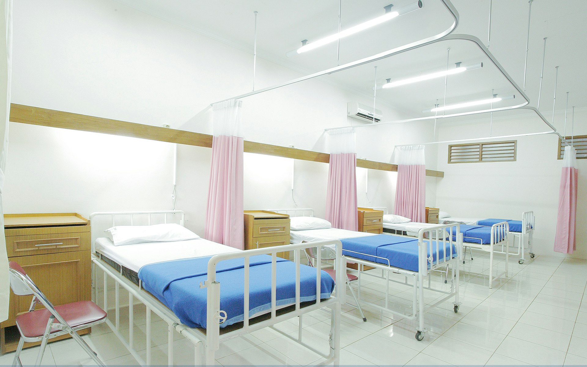 A clean and modern hospital ward with multiple beds, each equipped with blue bedding and separated by pink curtains for privacy.