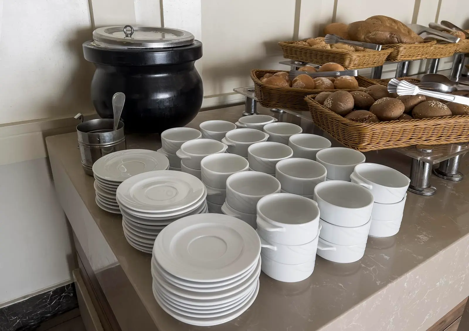 A display of white crockery, cups, and a basket of bread on a buffet table.