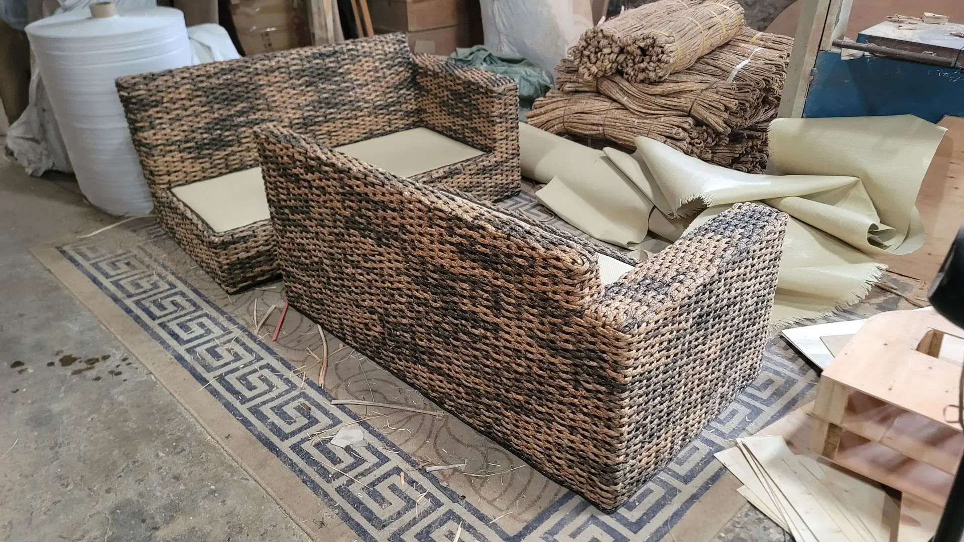 Rattan garden furniture set arranged in a workshop setting, featuring two modular seating pieces with beige cushions, surrounded by materials and other furniture components.