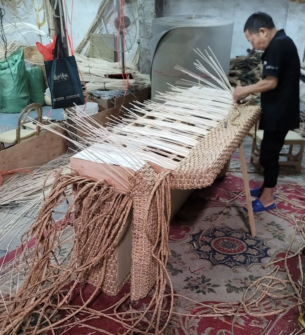 Craftsman weaving a rattan garden furniture set, focusing on detailed craftsmanship with natural materials and tools, in a workshop environment.