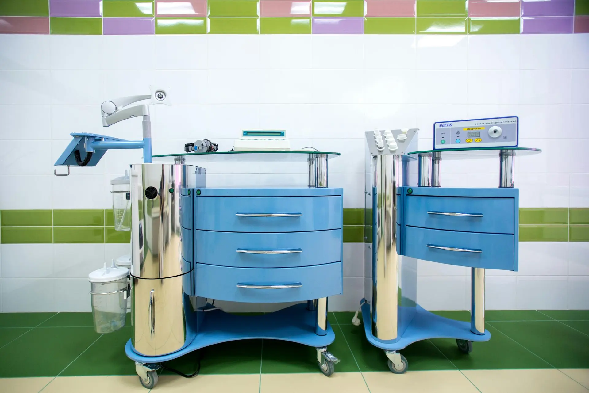 Close-up of two modern medical trolleys in blue, featuring storage drawers and various medical equipment on glass tops, set against a colorful tiled background.