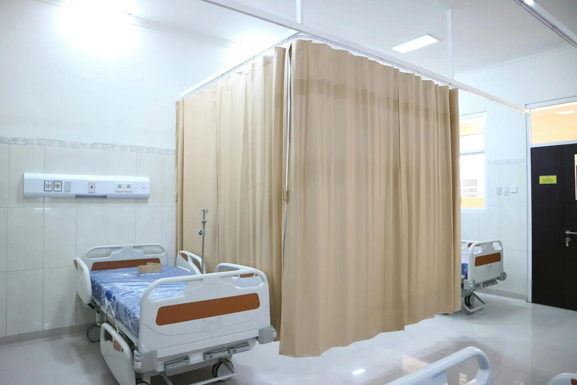 A hospital room featuring two patient beds separated by beige curtains, with medical equipment mounted on the wall and a door leading outside.
