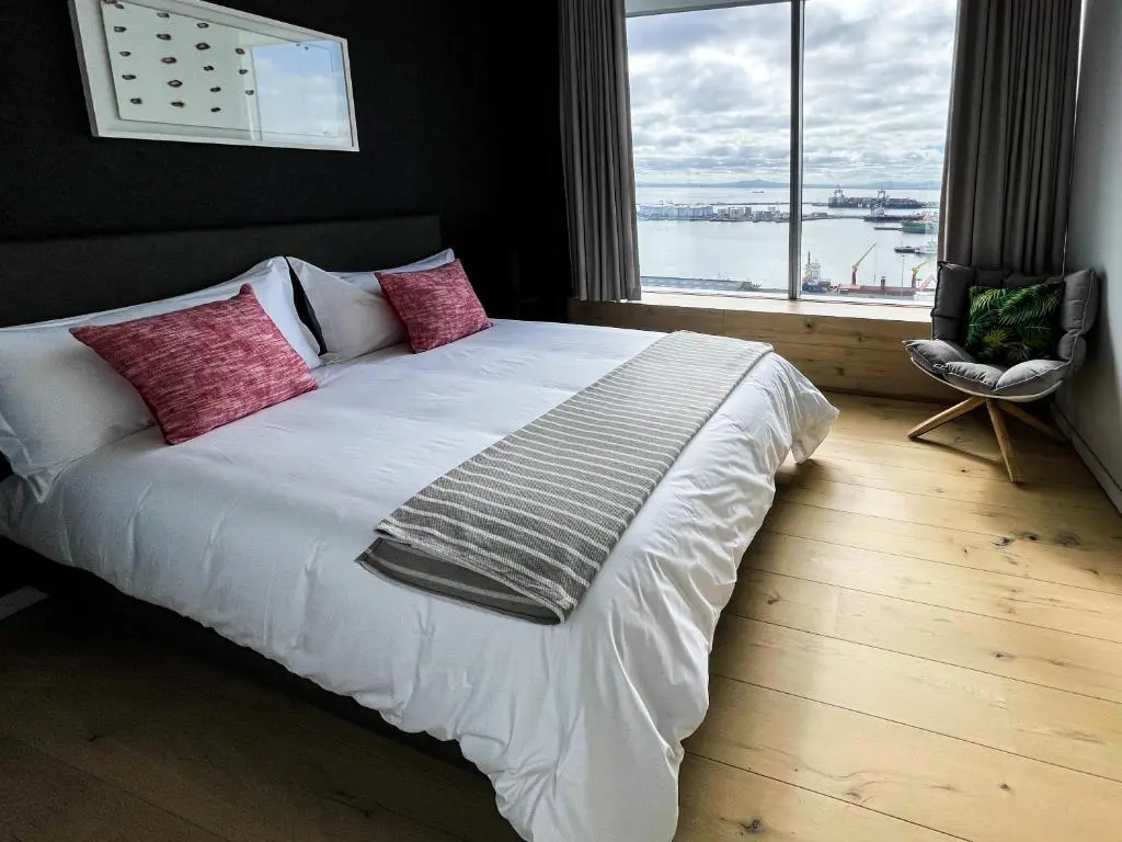 A modern bedroom featuring a stylish bed with red pillows, a textured throw, and a window view of the harbor in Cape Town