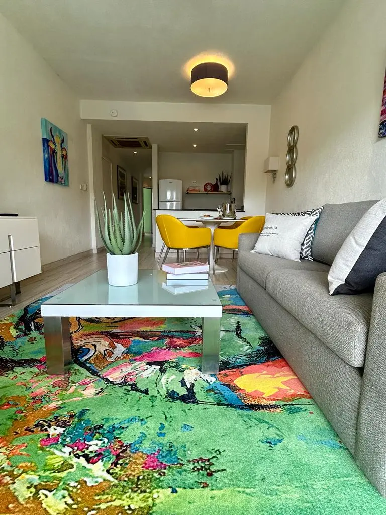 Stylish living area at The Rockley featuring a modern sofa, a colorful area rug, and a dining space in the background with yellow chairs and a welcoming atmosphere.
