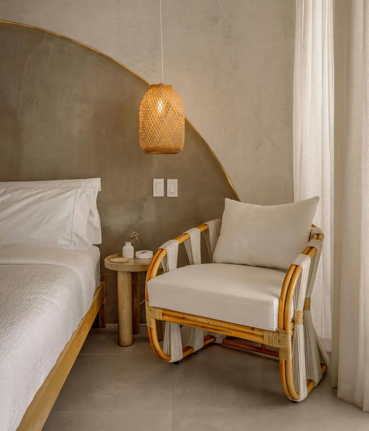 A rattan armchair with white cushions sits next to a bed with white bedding, a small round side table, and a hanging woven pendant lamp in a neutral-toned room—ideal inspiration for retirement home furniture.