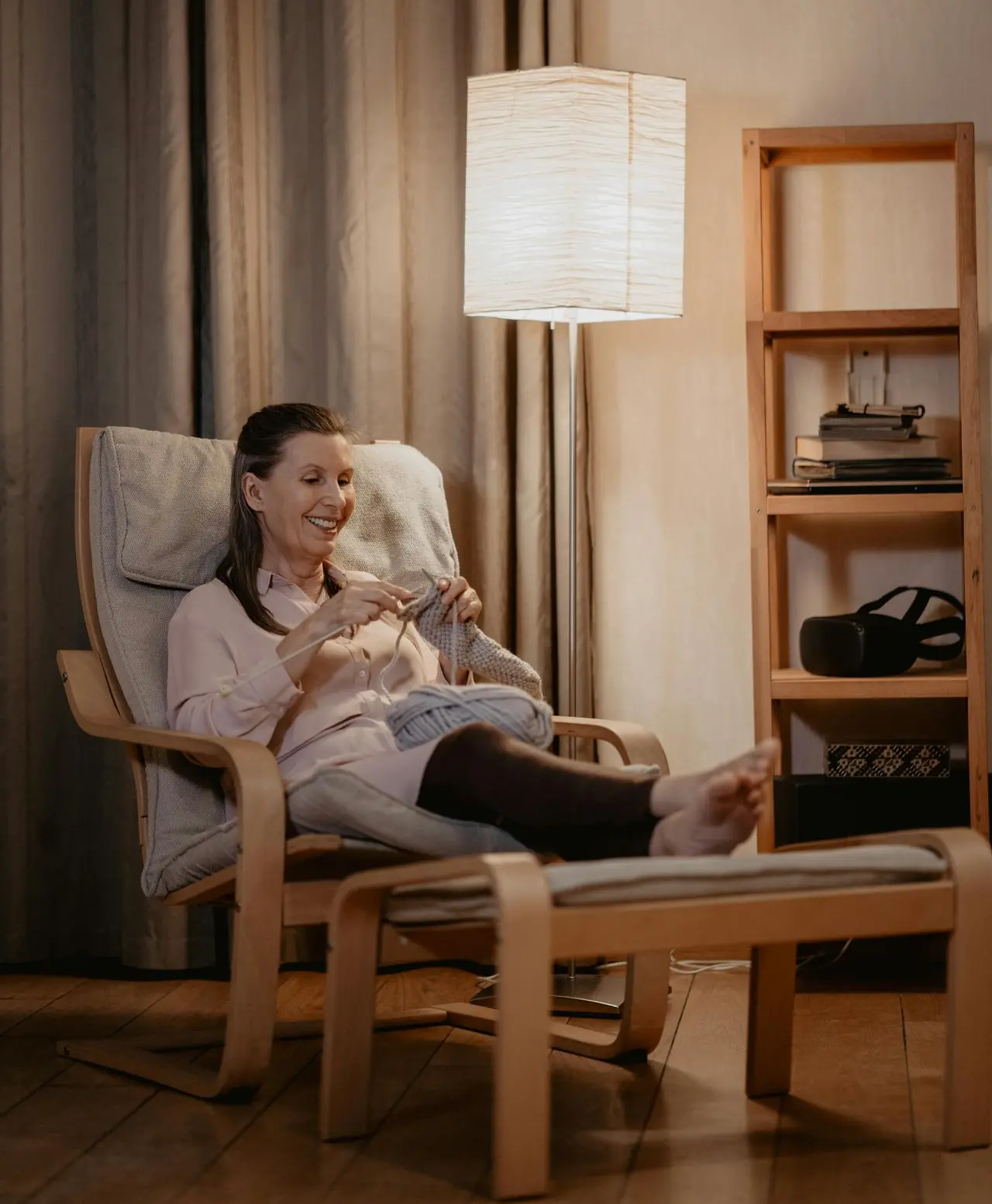 A woman sits in a cushioned chair with her feet up on an ottoman, knitting and smiling. A floor lamp and wooden shelves are in the background.