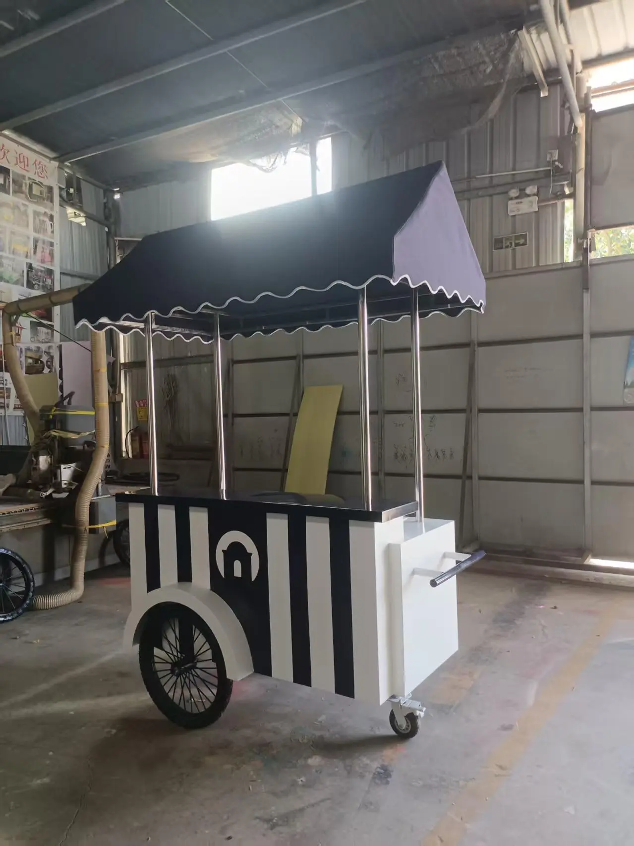 ecorative Vintage-Style Cart with Canopy – Black and White Striped Stand and Large Wheel A black and white striped cart with a canopy and two wheels is positioned inside a workshop with metal walls and windows.