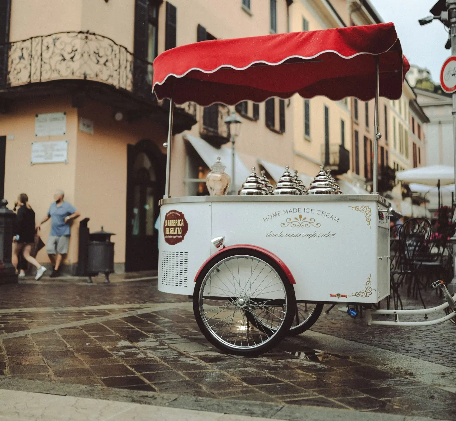 Ice Cream Cart on a Cobblestone Street An ice cream cart with a red canopy stands on a cobblestone street in front of buildings, with people and outdoor seating in the background.