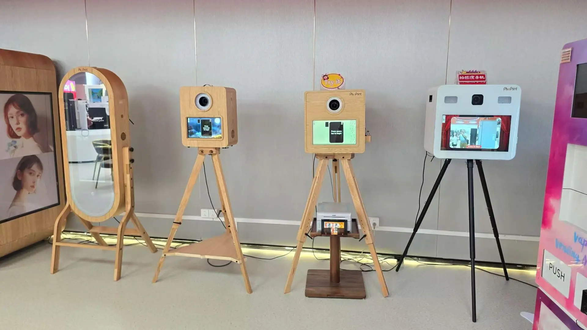 Four photo booth machines, three with wooden casings on tripods and one white machine on black legs, are displayed side by side in a modern indoor setting.