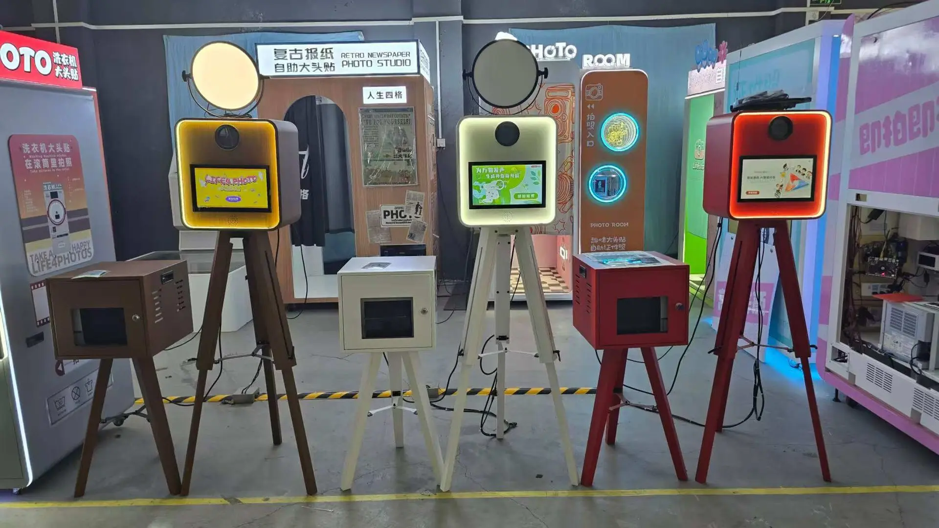 Five freestanding digital photo booths are displayed indoors, each with a screen, circular light on top, and various colored stands. Signs with Chinese text are visible in the background.