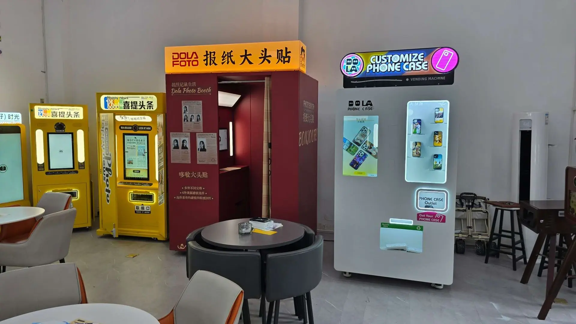 A photo booth, two yellow self-service kiosks, and a phone case customization vending machine are positioned against a wall in a modern indoor space with tables and chairs.