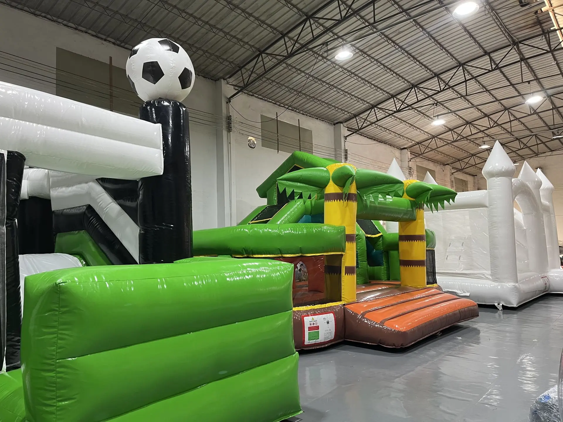 Indoor Inflatable Obstacle Course - Jungle & Soccer Ball Theme An indoor inflatable obstacle course with a green palm tree-themed section, brown steps, and a large black-and-white soccer ball on top of a white column; additional white inflatable structures are visible in the background.
