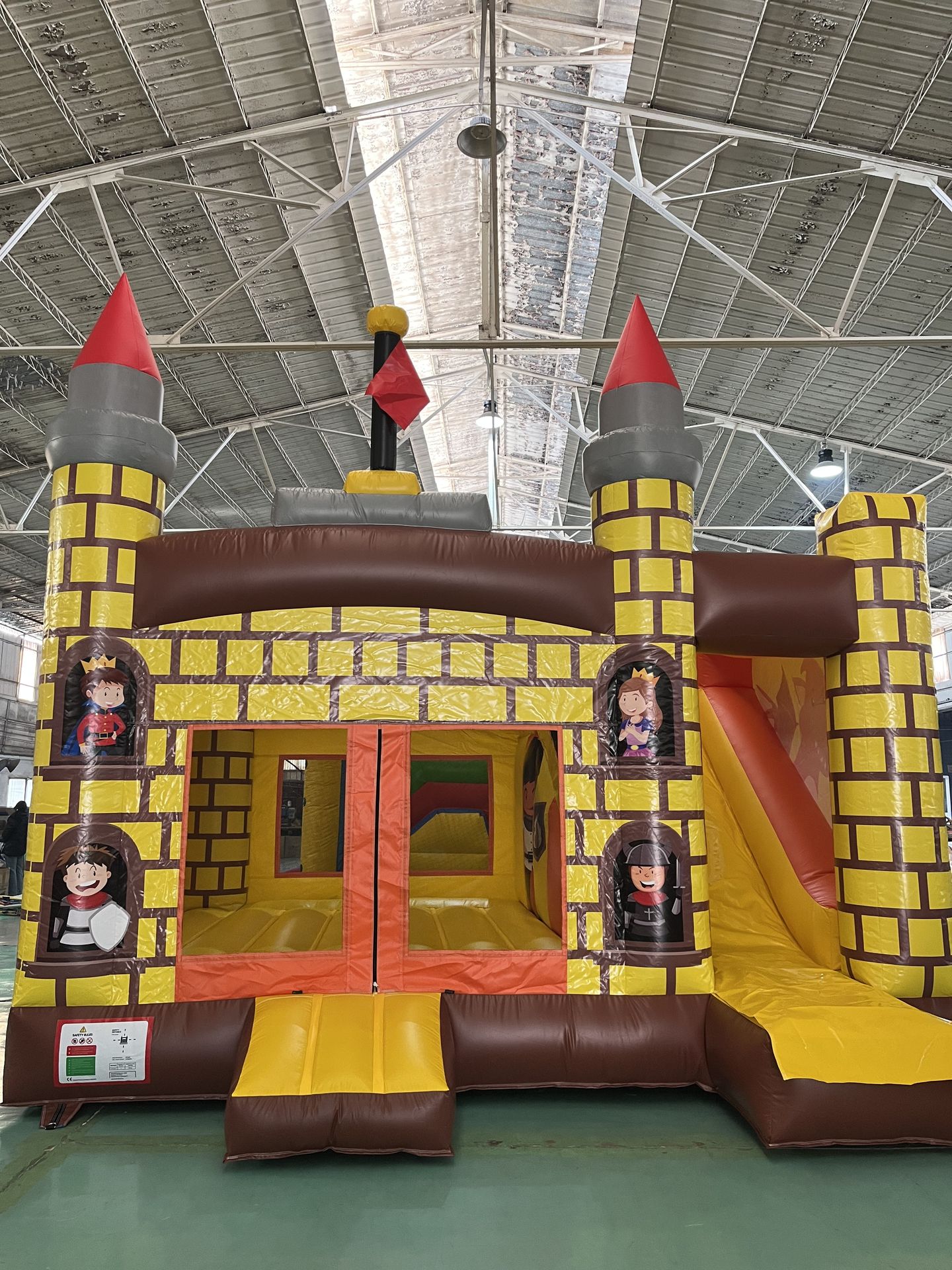 Inflatable Castle Playground with Slid Colorful inflatable castle with yellow brick-pattern walls, two round towers topped with red cones, a brown and yellow ramp, and a slide on the right side