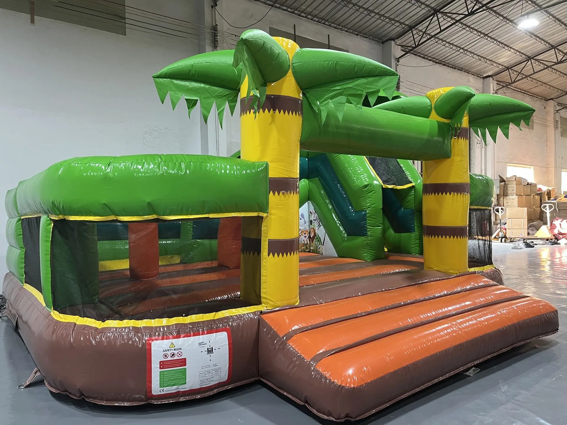 Inflatable Jungle Obstacle Course with Palm Trees and Slide A large inflatable jungle-themed obstacle course inside a warehouse, featuring green palm-tree arches, yellow and brown pillars, a brown and orange ramp, and a green slide in the background.