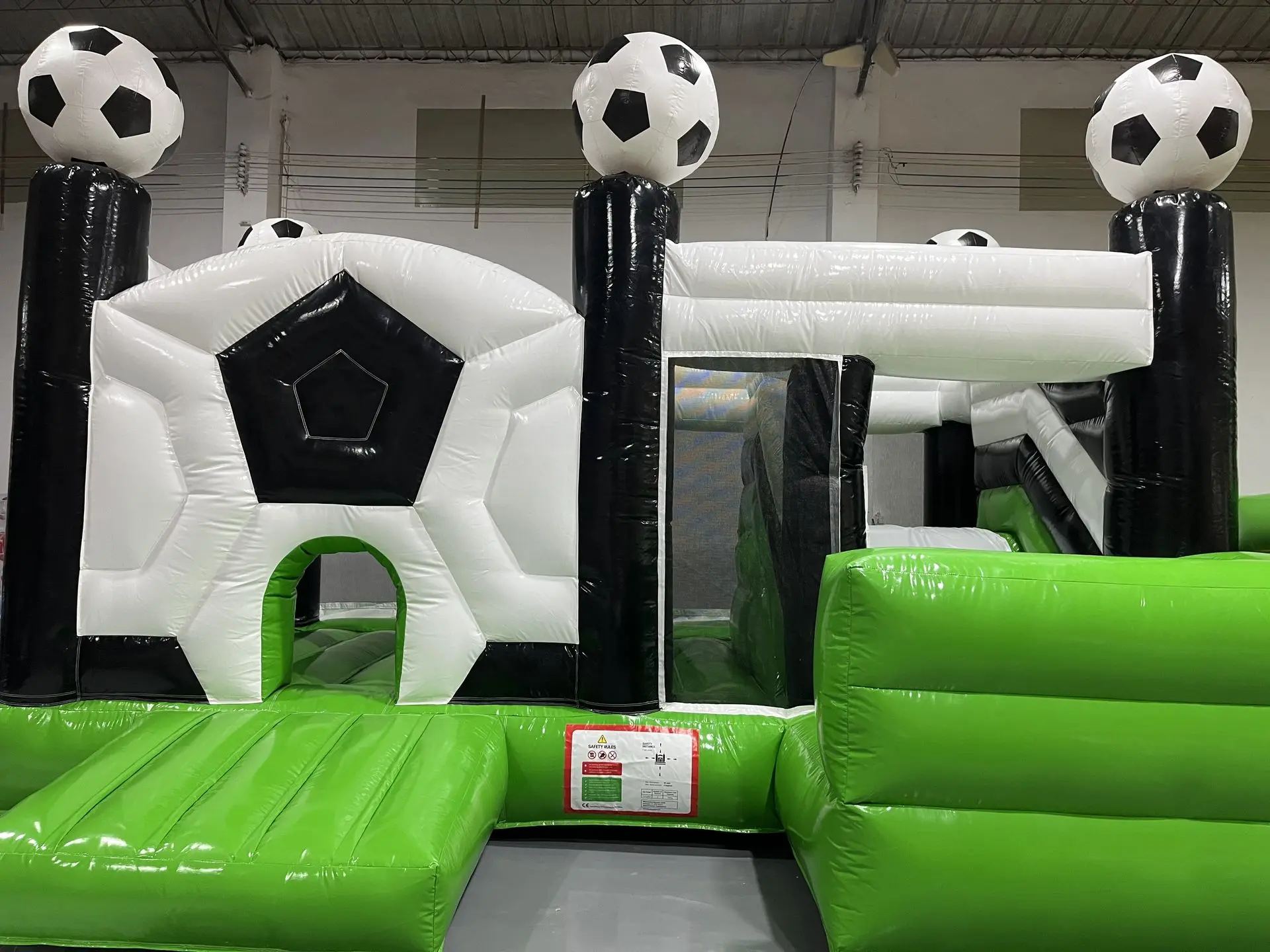 Inflatable Soccer-Themed Obstacle Course with Ball-Topped Towers inflatable soccer-themed obstacle course with black and white goal-like arches, large soccer ball toppers on tall black columns, green inflatable platforms, and a white tube entrance.