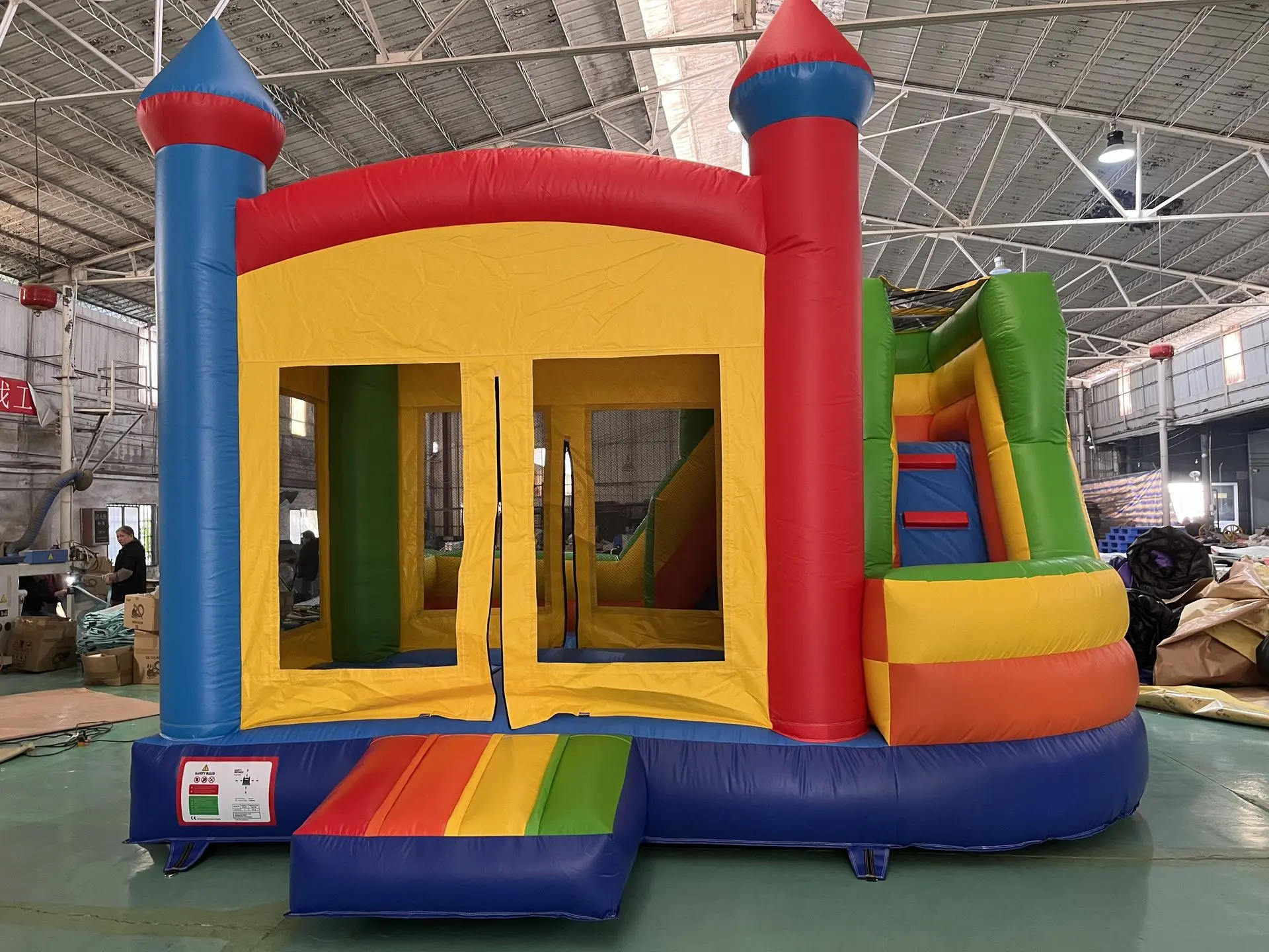 Inflatable Castle Bounce House with Rainbow Ramp and Slid Colorful inflatable bounce house with a yellow central panel, blue and red towers at the corners, a small blue ramp with rainbow stripes