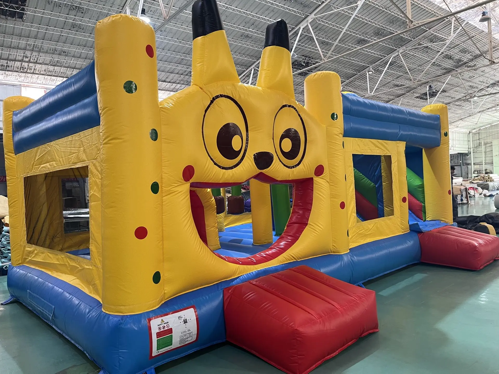 Inflatable Castle Playground with Cartoon Face Entrance A bright inflatable castle with a cheerful yellow façade, large cartoon eyes, red mouth opening as the entrance, and blue and yellow towers; a red inflatable step sits at the front