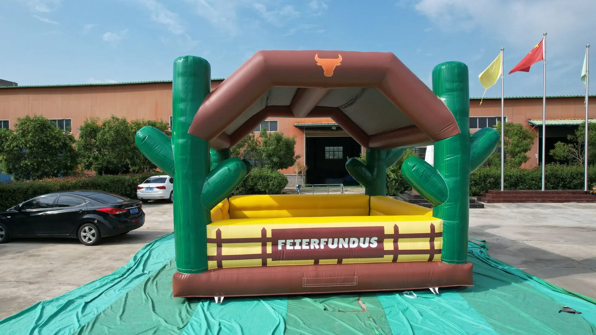 Outdoor Inflatable Playground with Cowboy Theme and Fiesta Roof Inflatable rodeo-style arena with a brown roof, green posts shaped like cacti, and a yellow rectangular pit labeled “FEIERFUNDUS” inside a large outdoor setup on a teal ground