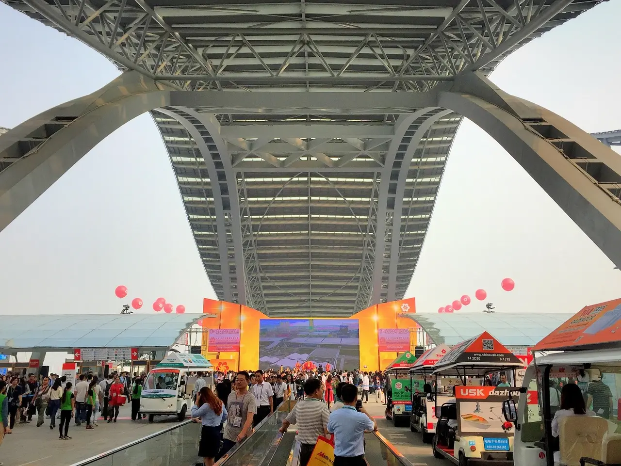 Crowded Canton Fair entrance under a large steel archway with orange stage, booths, and red balloons in the background