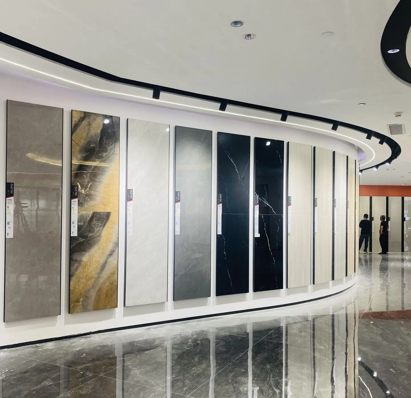 Tile samples on curved showroom wall A curved showroom wall with a row of large tile samples in various colors and patterns, including beige, gray, and black marble, reflected on a polished floor.