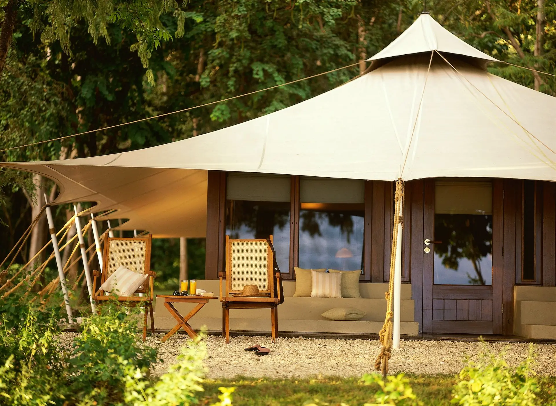 Outdoor tented shade structure with beige fabric, wooden seating, and glass doors in a garden setting