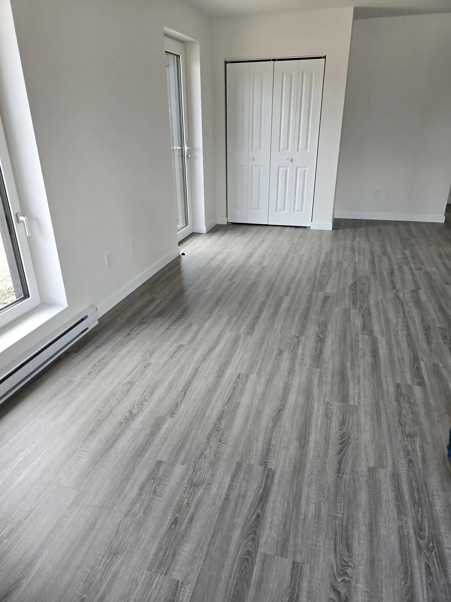 Gray Weathered Wood-Look SPC Flooring in Modern Residential Space Newly installed SPC or luxury vinyl plank flooring with a gray weathered wood-grain look in a bright, empty room with white walls, a white bi-fold closet, and white-trimmed windows.