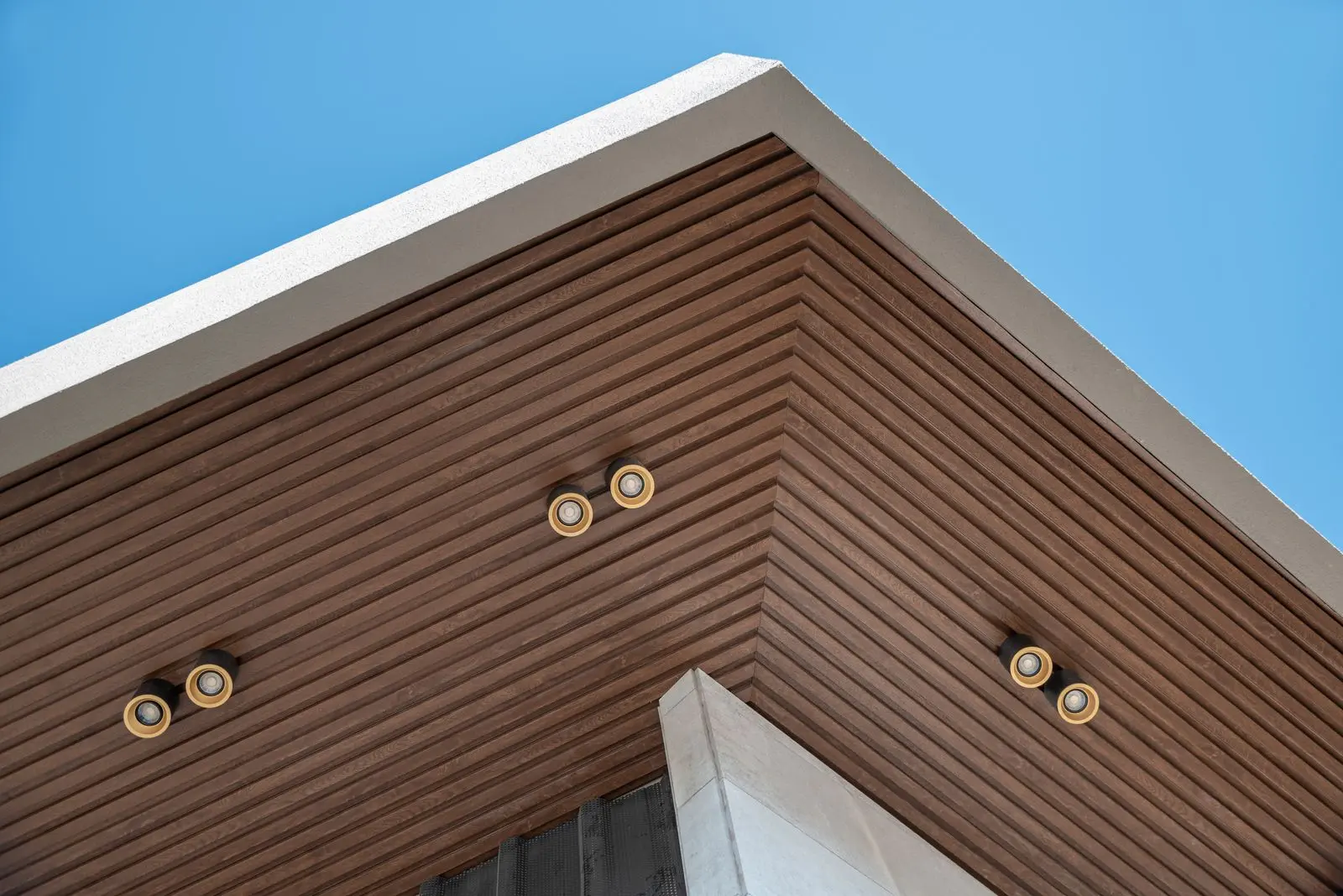 WPC Ceilings: Wood-look Aesthetics with Water and Termite Resistance for Indoor and Semi-Outdoor Spaces Close-up of a building’s exterior corner with wood-grain WPC ceiling panels, showing natural brown slats and outdoor lighting fixtures against a blue sky.