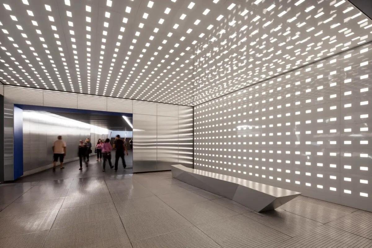 Contemporary metal-ceiling transit hall with illuminated perforations A modern subway or transit station hallway with a perforated metal ceiling featuring evenly spaced square cutouts that glow with light, plus a sleek metal bench along the wall and a bright, reflective floor.