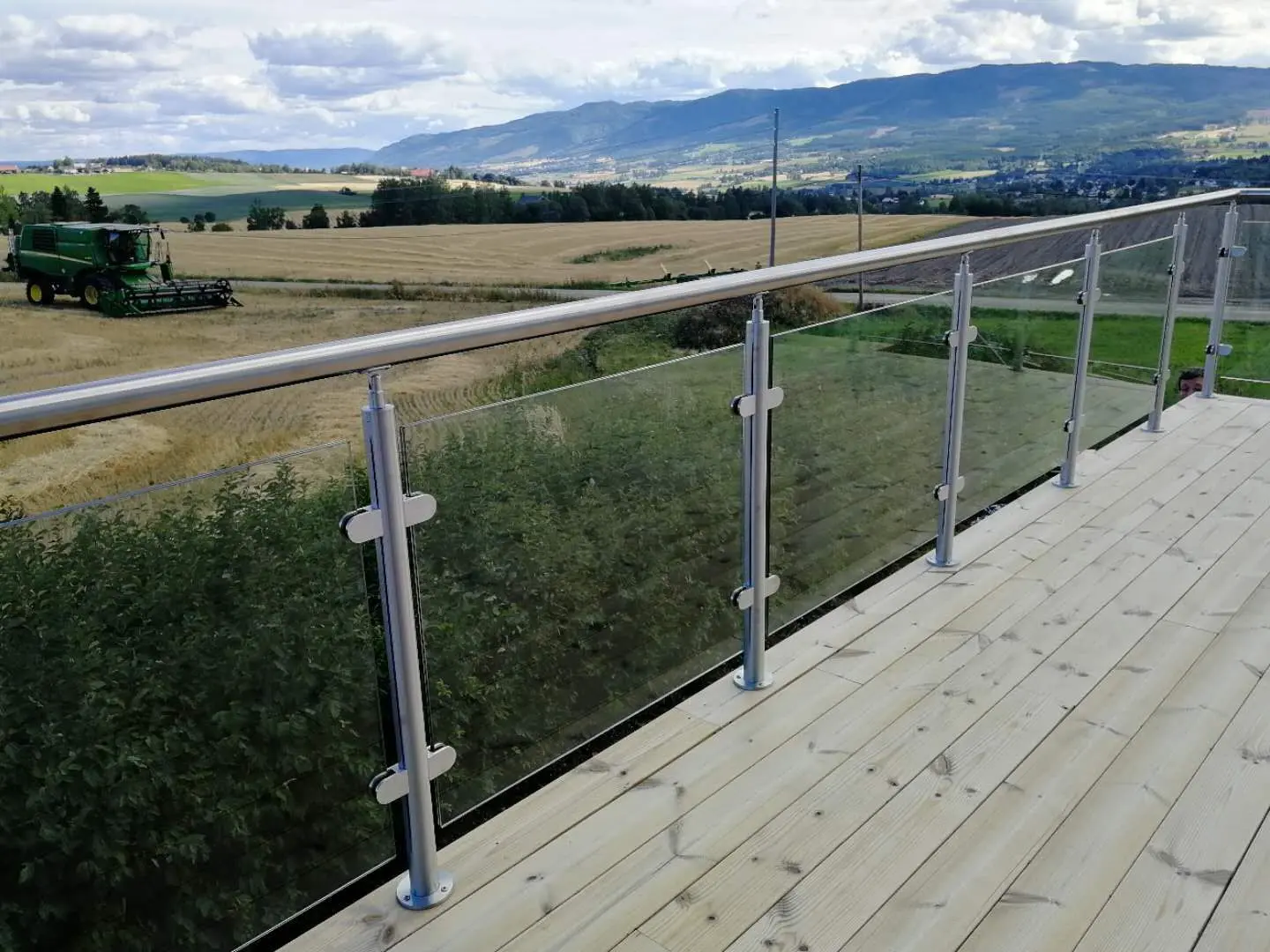 Glass railing on a wooden balcony with stainless steel posts, overlooking a rural landscape with fields and hills.