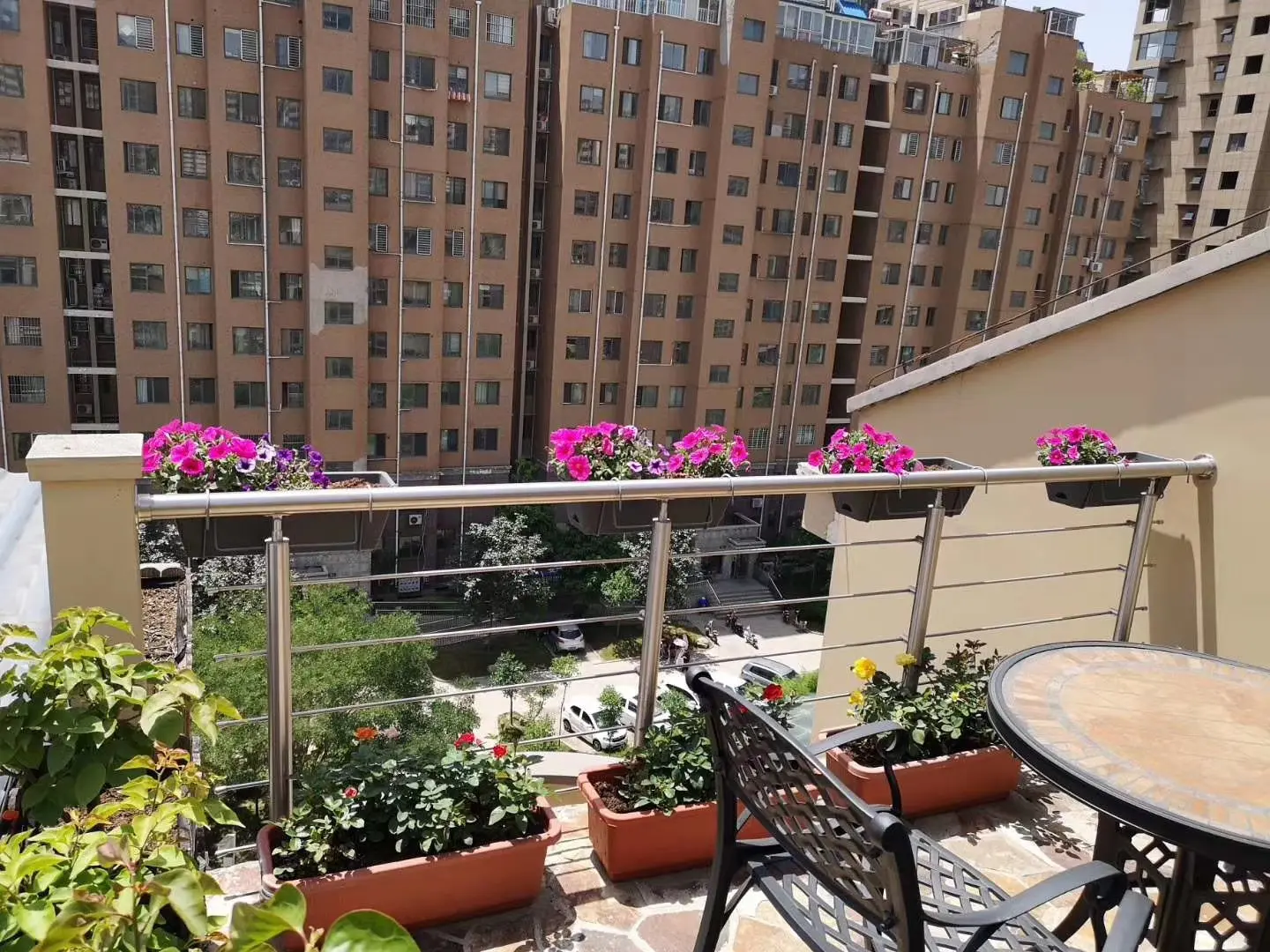 Rooftop balcony with stainless steel railing, planters with pink flowers, and outdoor seating overlooking an urban residential complex.