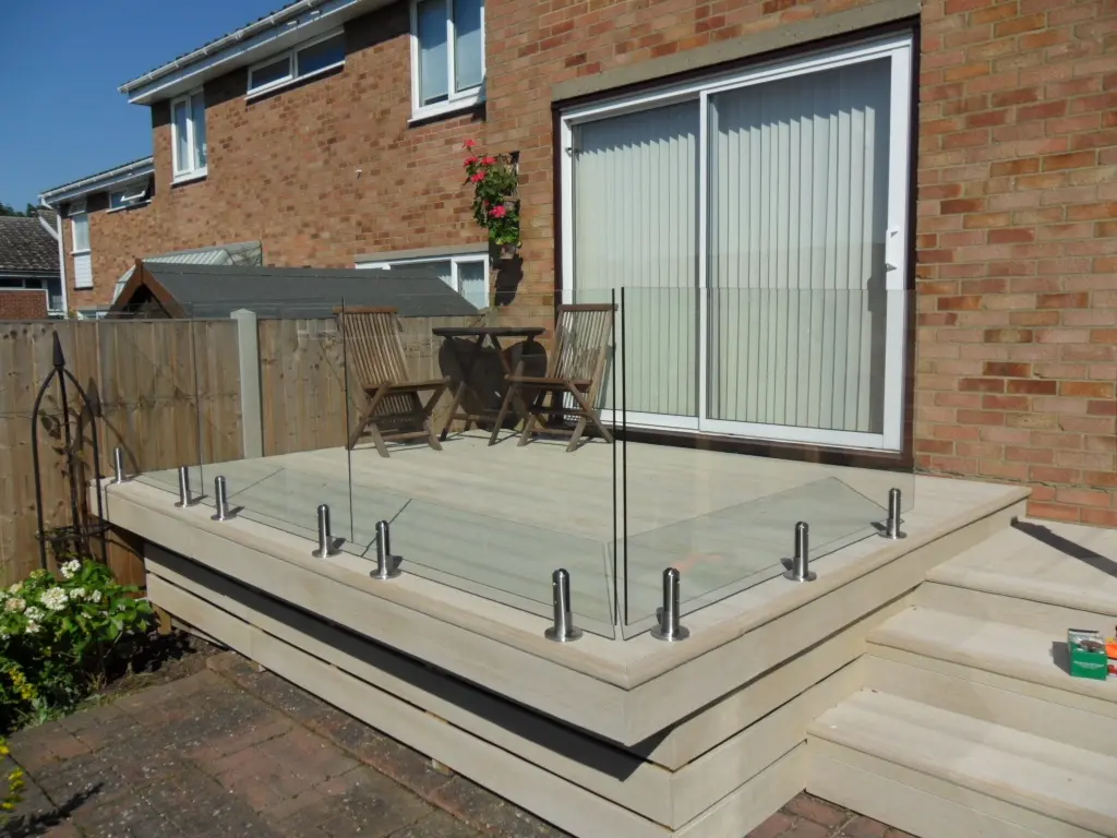 Residential deck with glass railing installed around the upper level, wooden steps leading down, and a small outdoor seating area against a brick house.
