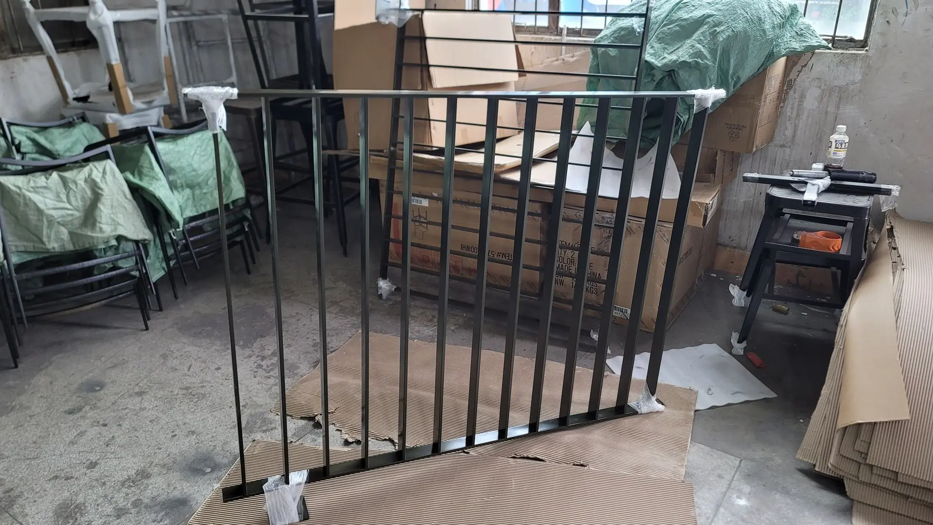 Metal railing being assembled in a workshop, with cardboard and work tools around, wrapped joints at the base.