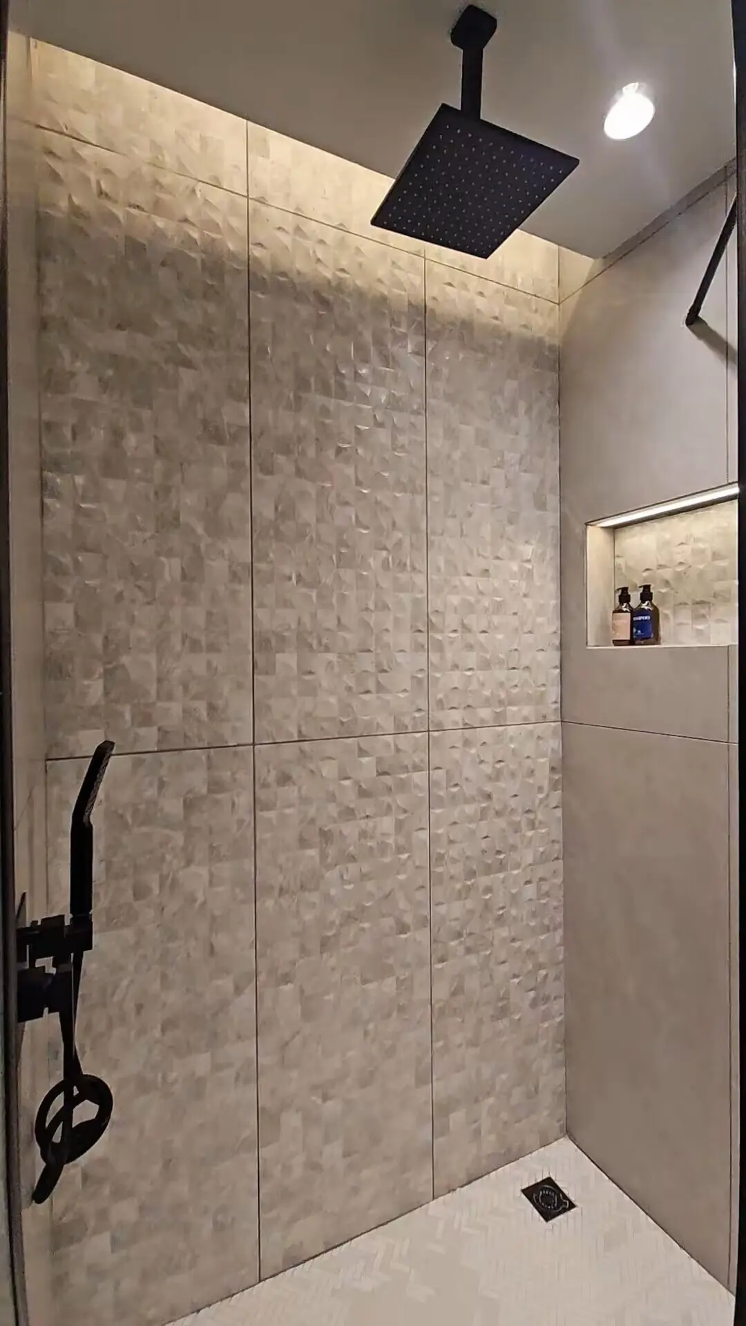 Rainfall Shower Fixtures - GZS Tiling and Plumbing OS&E Modern hotel shower with textured white tile, black matte rainfall showerhead, and recessed shelf, demonstrating GZS's sourcing of high-end fixtures and materials.