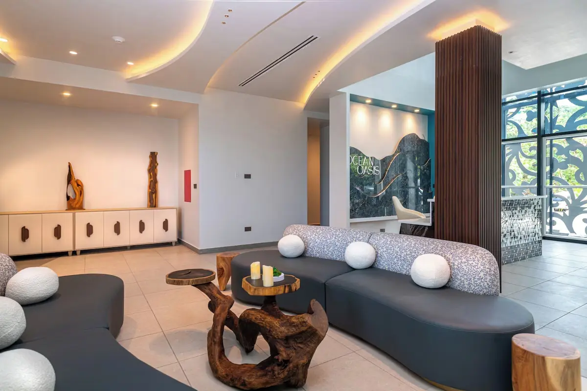 Custom Lounge Seating and Accent Tables - GZS Sourced FF&E Modern hotel lounge area featuring curved grey sofas, unique natural wood tables, and a feature wall with the Ocean Oasis logo, sourced as FF&E by GZS.