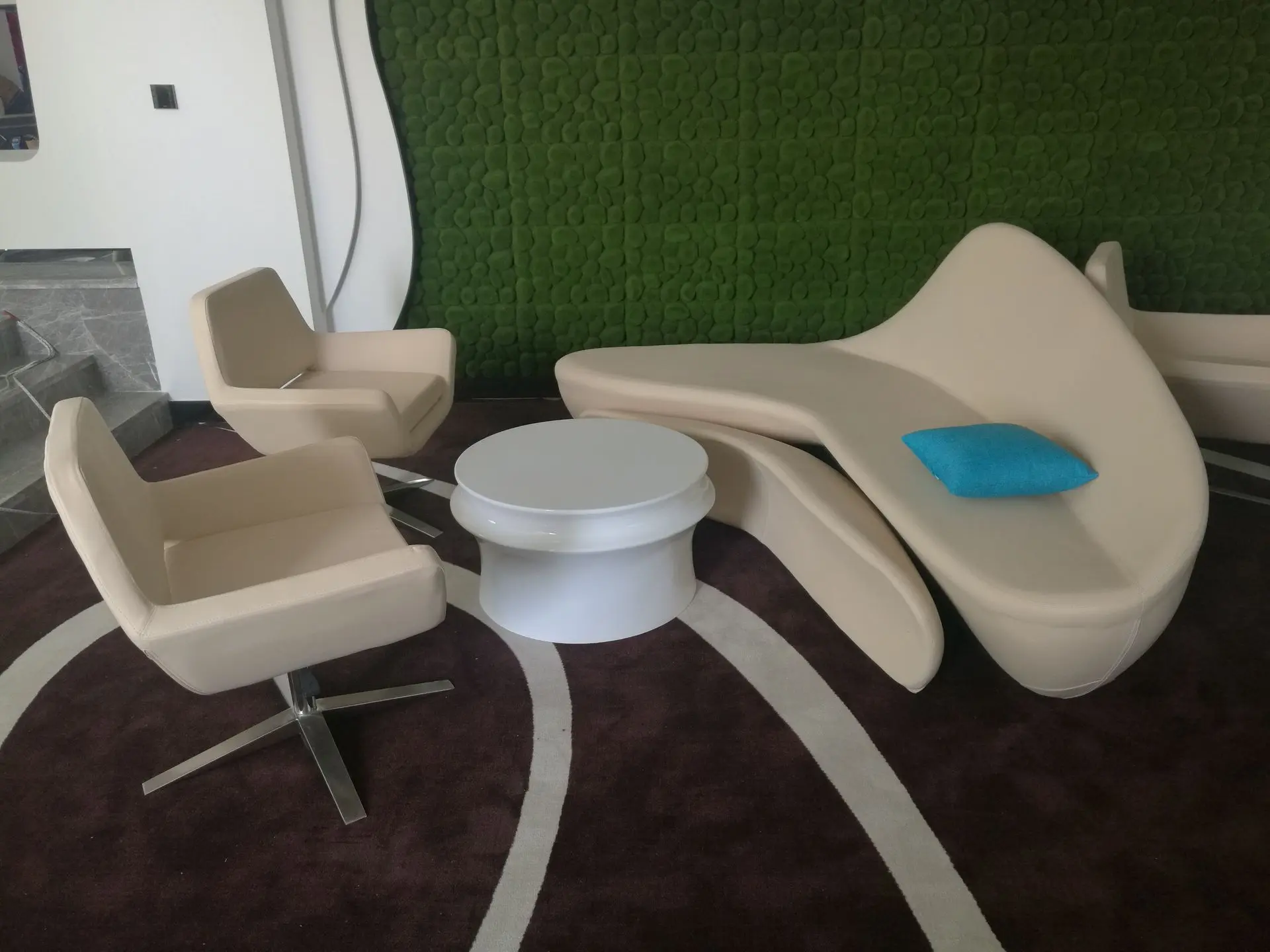 Fiberglass Molded Seating: Futuristic Loose Furniture China Sourcing Modern lounge area featuring futuristic loose furniture china seating, including a curved, sculptural beige fiberglass sofa with a blue cushion, two swivel chairs, and a round white table, set against a green moss wall.