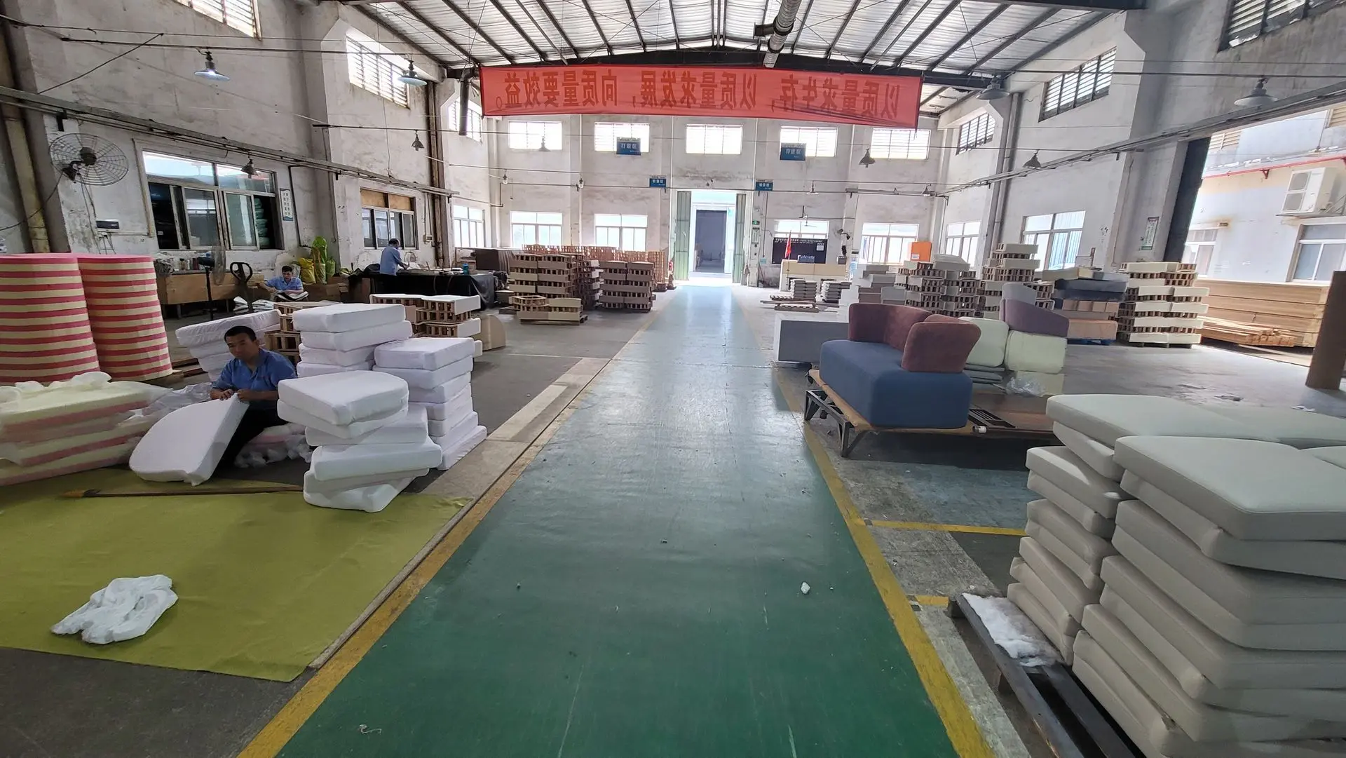 Interior view of a loose furniture china factory floor showing large stacks of foam, upholstered cushions, and workers preparing components for sofa and seating production.