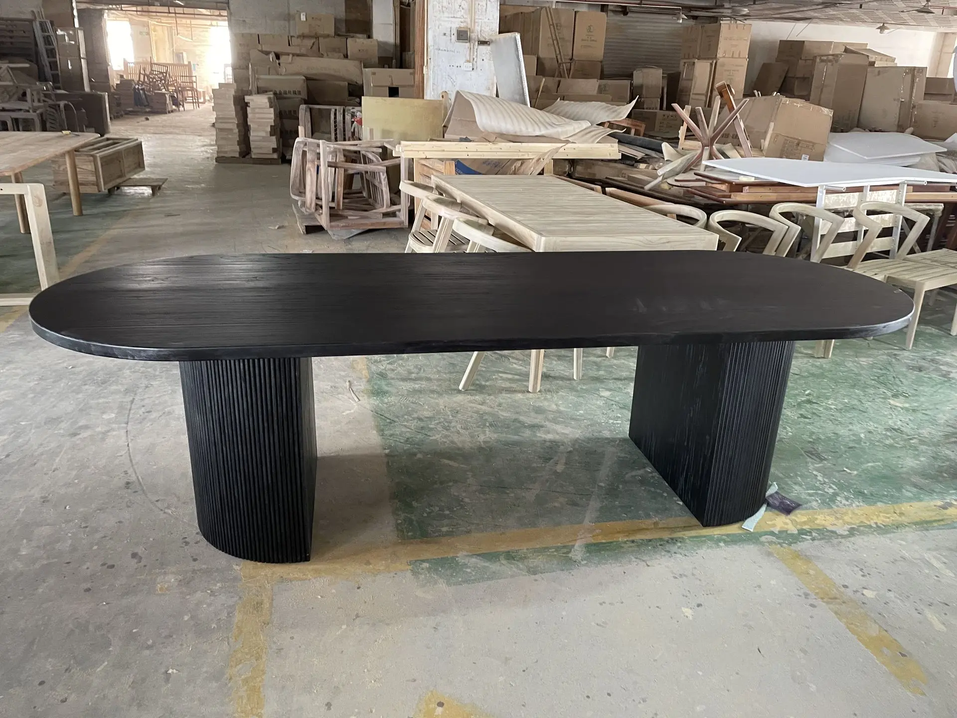 Black Solid Teak Dining Table: Custom Oval Loose Furniture China Oval dining table with a black textured finish, supported by two fluted cylindrical wood bases, showcasing modern large-scale loose furniture china manufacturing in a workshop.