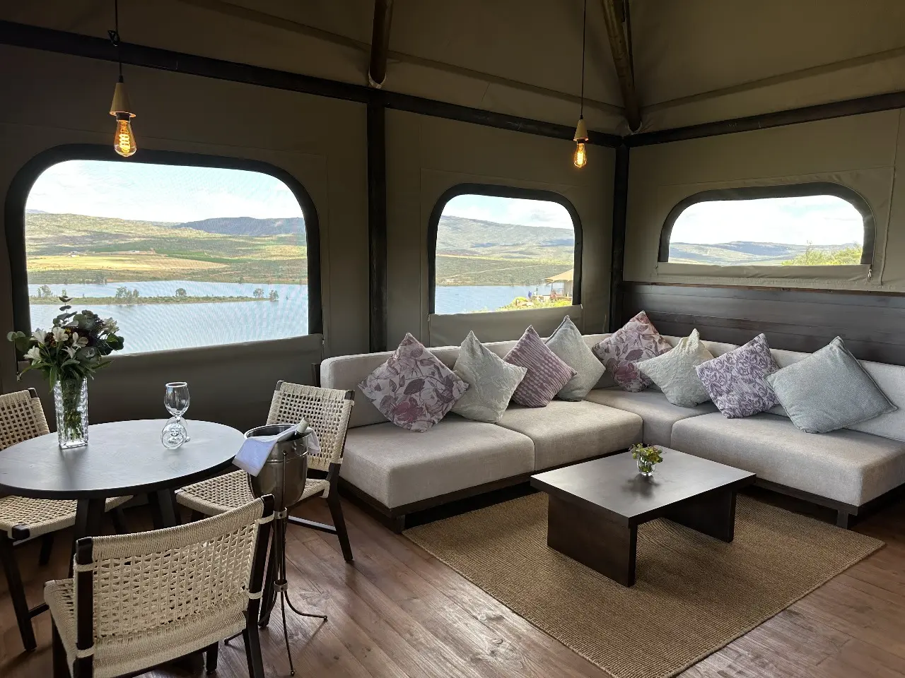 Yamu tent villa interior with woven dining chairs, fabric sectional sofa, wooden flooring, and panoramic lake and mountain views.