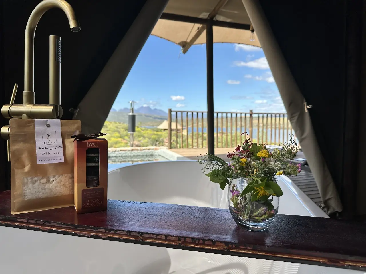 Yamu bathtub area with brass faucet, flowers, and open canopy view of the natural landscape.